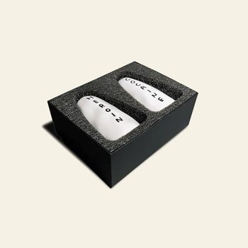 The Shrig Shop Heroin and Cocaine Porcelain Shakers by Shrig Shop are two white, oval shakers labeled with black text and numbers, housed in a black foam-padded box—a collectible set inspired by David Shrigley.