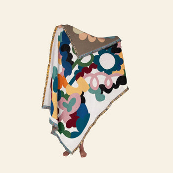 A person stands with only their feet showing, holding up the Slowdown Studio Alessi Throw—an abstract, bold-patterned blanket made from recycled cotton—contrasting against a plain light beige background.