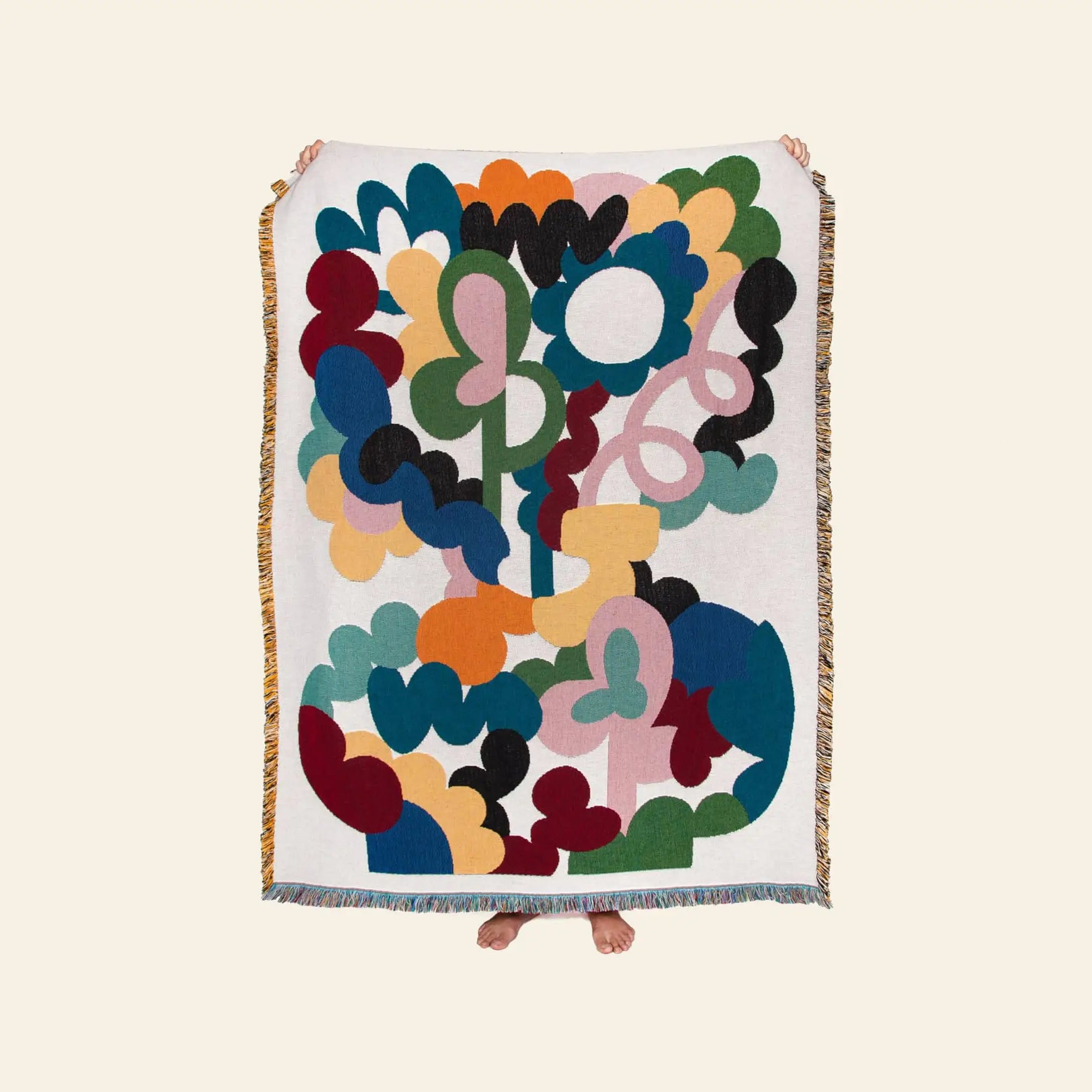 A person holds up the Slowdown Studio Alessi Throw, a sustainable blanket made from recycled cotton with colorful abstract designs and fringe edges. Only their hands and feet are visible behind the eco-friendly Slowdown Studio piece.
