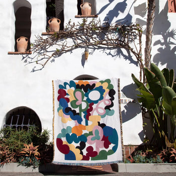 The Slowdown Studio Alessi Throw, a colorful abstract blanket made from recycled cotton, is displayed in front of a white stucco building with arched windows, terracotta pots, and lush green plants.