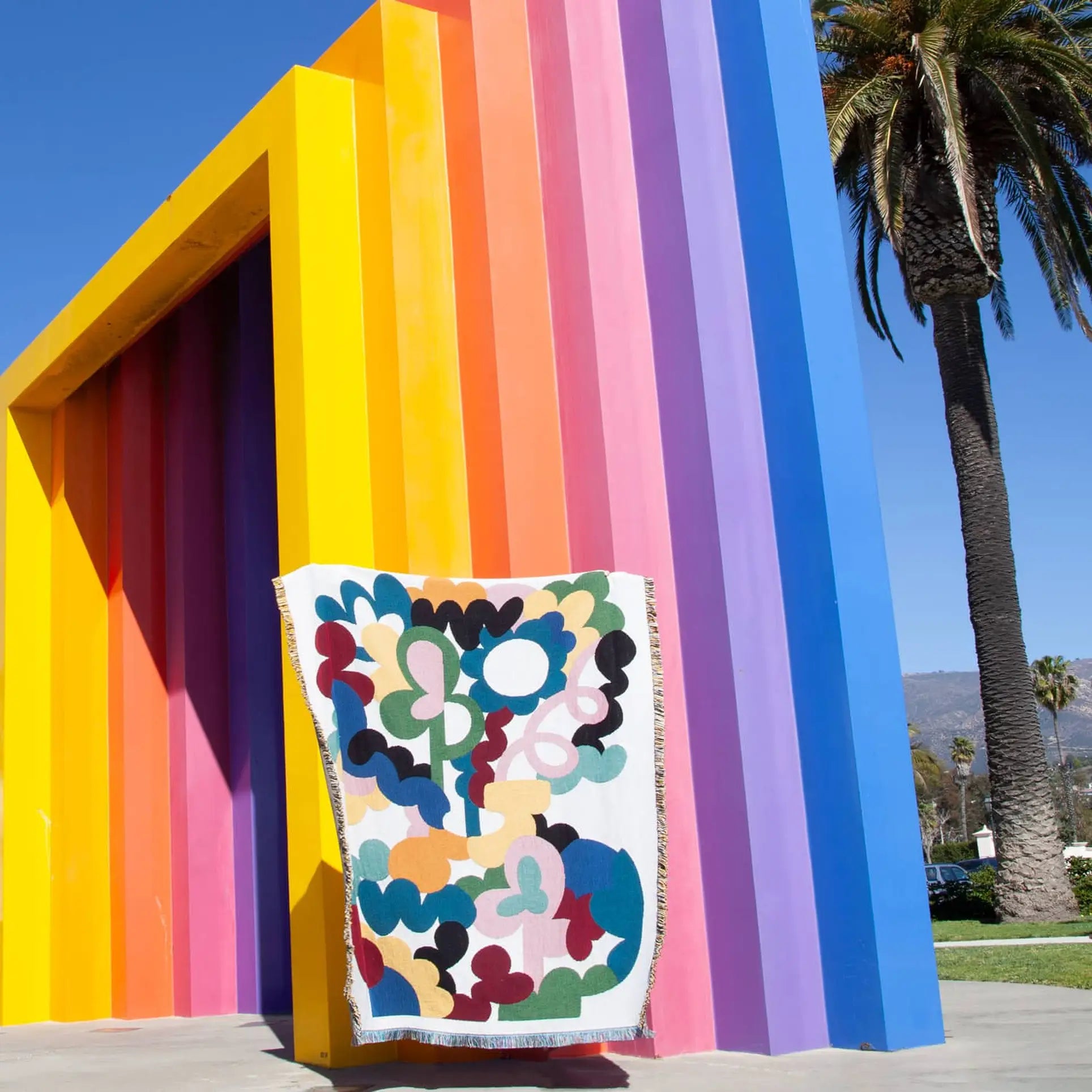 The Slowdown Studio Alessi Throw, made from recycled cotton by Slowdown Studio, drapes over a bold striped sculpture in yellow, orange, pink, and purple. Tall palm trees and a blue sky complete this eco-friendly scene.