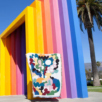 The Slowdown Studio Alessi Throw, made from recycled cotton by Slowdown Studio, drapes over a bold striped sculpture in yellow, orange, pink, and purple. Tall palm trees and a blue sky complete this eco-friendly scene.