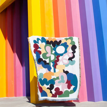 The Slowdown Studio Alessi Throw, featuring a colorful abstract pattern and made from recycled cotton, is displayed against a vibrant wall with vertical stripes—creating an eye-catching, artistic scene.
