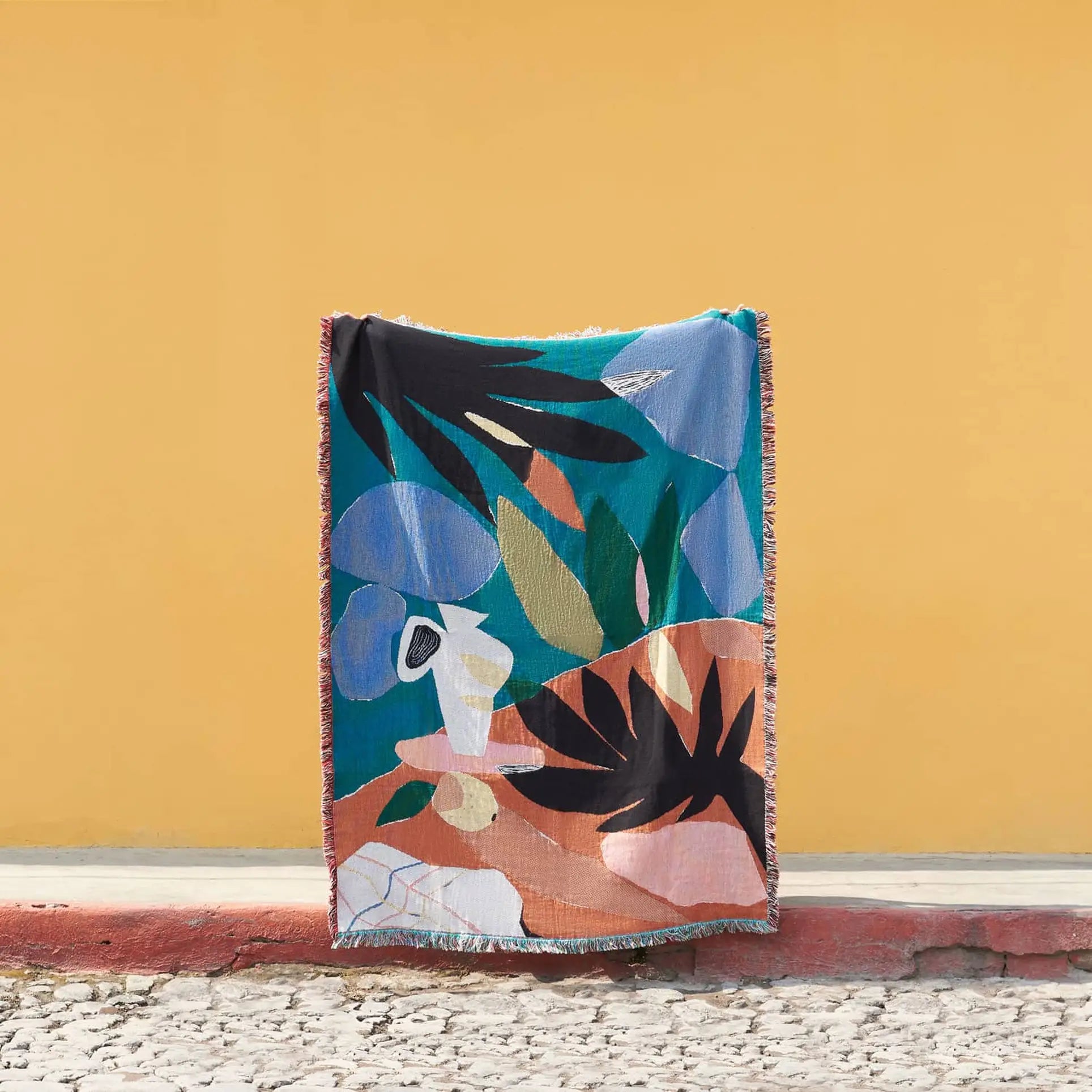 The Slowdown Studio Barbosa Throw, crafted from recycled cotton, stands upright against a yellow wall. This colorful blanket features bold abstract shapes in blue, green, orange, black, and white on cobblestone pavement.