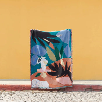The Slowdown Studio Barbosa Throw, crafted from recycled cotton, stands upright against a yellow wall. This colorful blanket features bold abstract shapes in blue, green, orange, black, and white on cobblestone pavement.