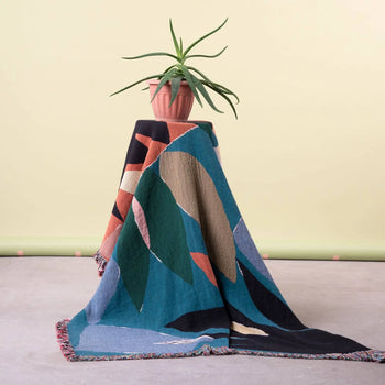A potted aloe vera plant sits on a small table draped with the Slowdown Studio Barbosa Throw by Slowdown Studio, made from recycled cotton and featuring colorful abstract patterns in blue, green, black, tan, and rust on light yellow.