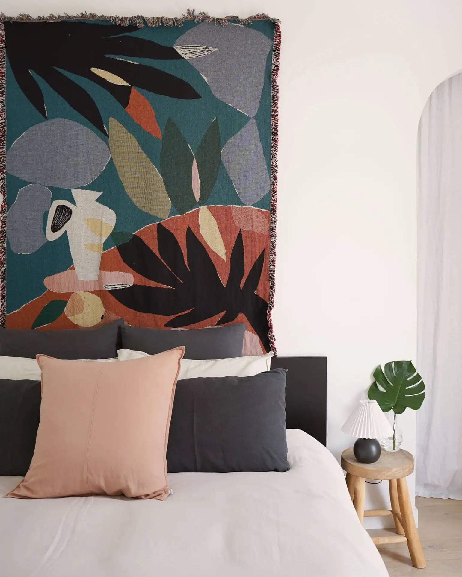 A modern bedroom displays a bed with black, white, and blush pillows under the Slowdown Studio Barbosa Throw. A wooden side table holds a white lamp, black vase, and green leaf. The wall is white with an arched opening.