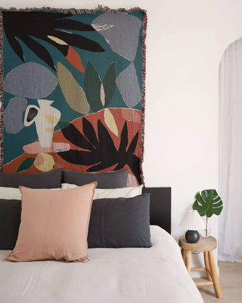 A modern bedroom displays a bed with black, white, and blush pillows under the Slowdown Studio Barbosa Throw. A wooden side table holds a white lamp, black vase, and green leaf. The wall is white with an arched opening.
