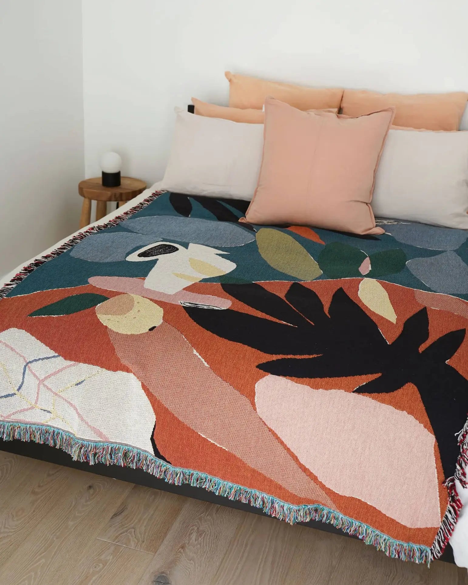 A neatly made bed with cream and peach pillows, a wooden nightstand with a lamp, and the Slowdown Studio Barbosa Throw—a colorful abstract blanket in red, pink, navy, green, and cream, crafted from recycled cotton by Slowdown Studio.