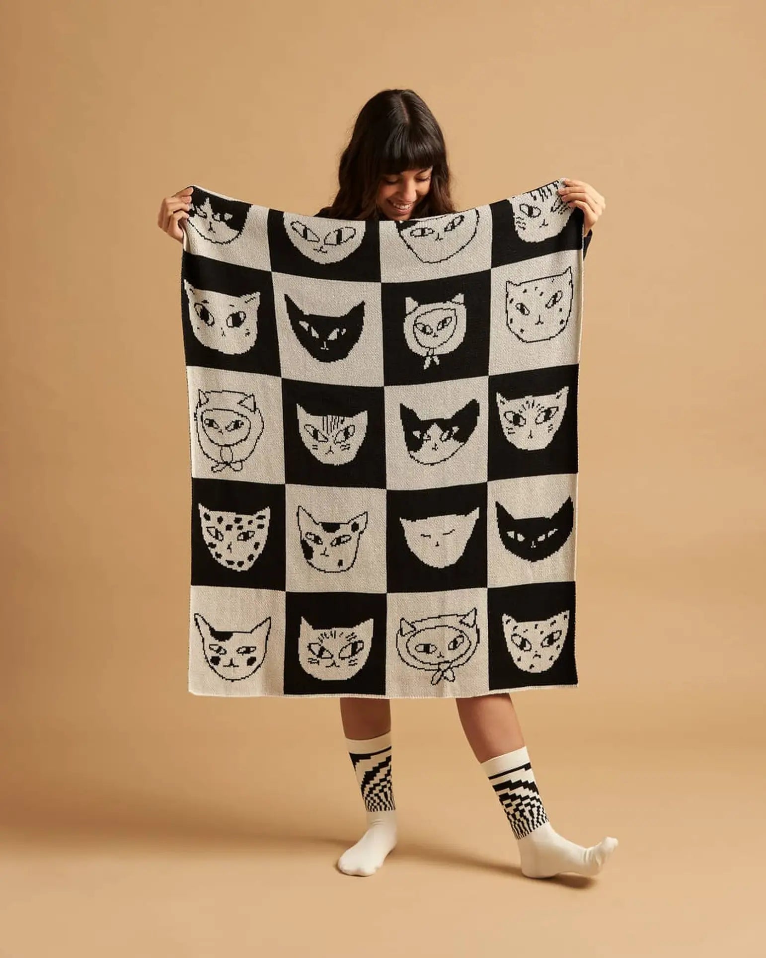 On a tan background, a person smiles while holding the Slowdown Studio Cat Bingo Mini Blanket—made by Slowdown Studio from recycled cotton blend, featuring a grid of black-and-white cat faces. They wear bold, mismatched socks.