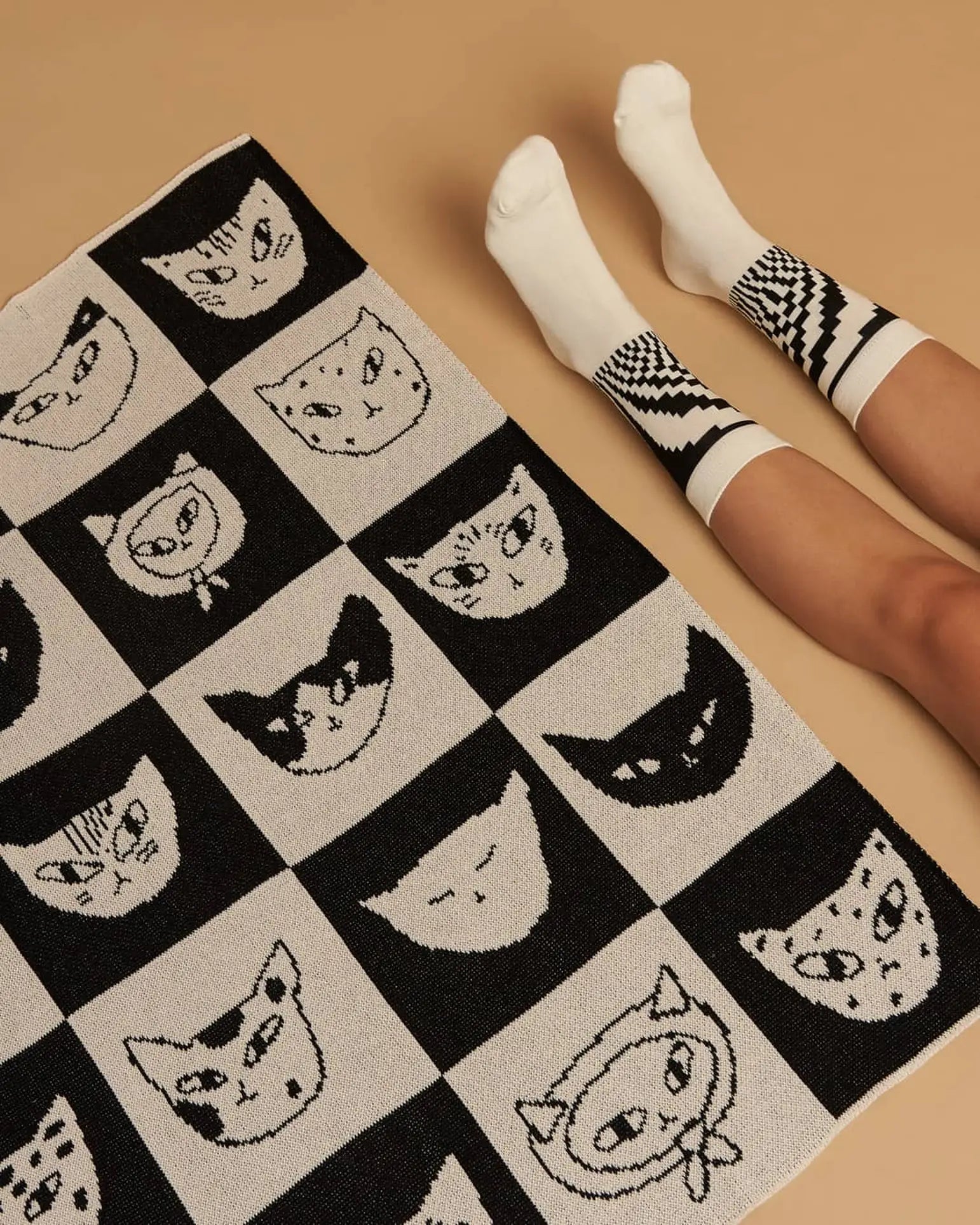 A person in white socks with black geometric patterns sits on a beige floor next to the Slowdown Studio Cat Bingo Mini Blanket by Slowdown Studio, featuring illustrated cat faces in a playful grid and made from a recycled cotton blend.