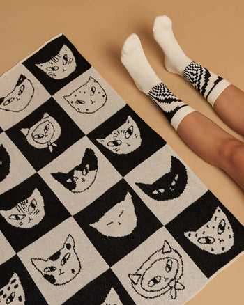 A person in white socks with black geometric patterns sits on a beige floor next to the Slowdown Studio Cat Bingo Mini Blanket by Slowdown Studio, featuring illustrated cat faces in a playful grid and made from a recycled cotton blend.