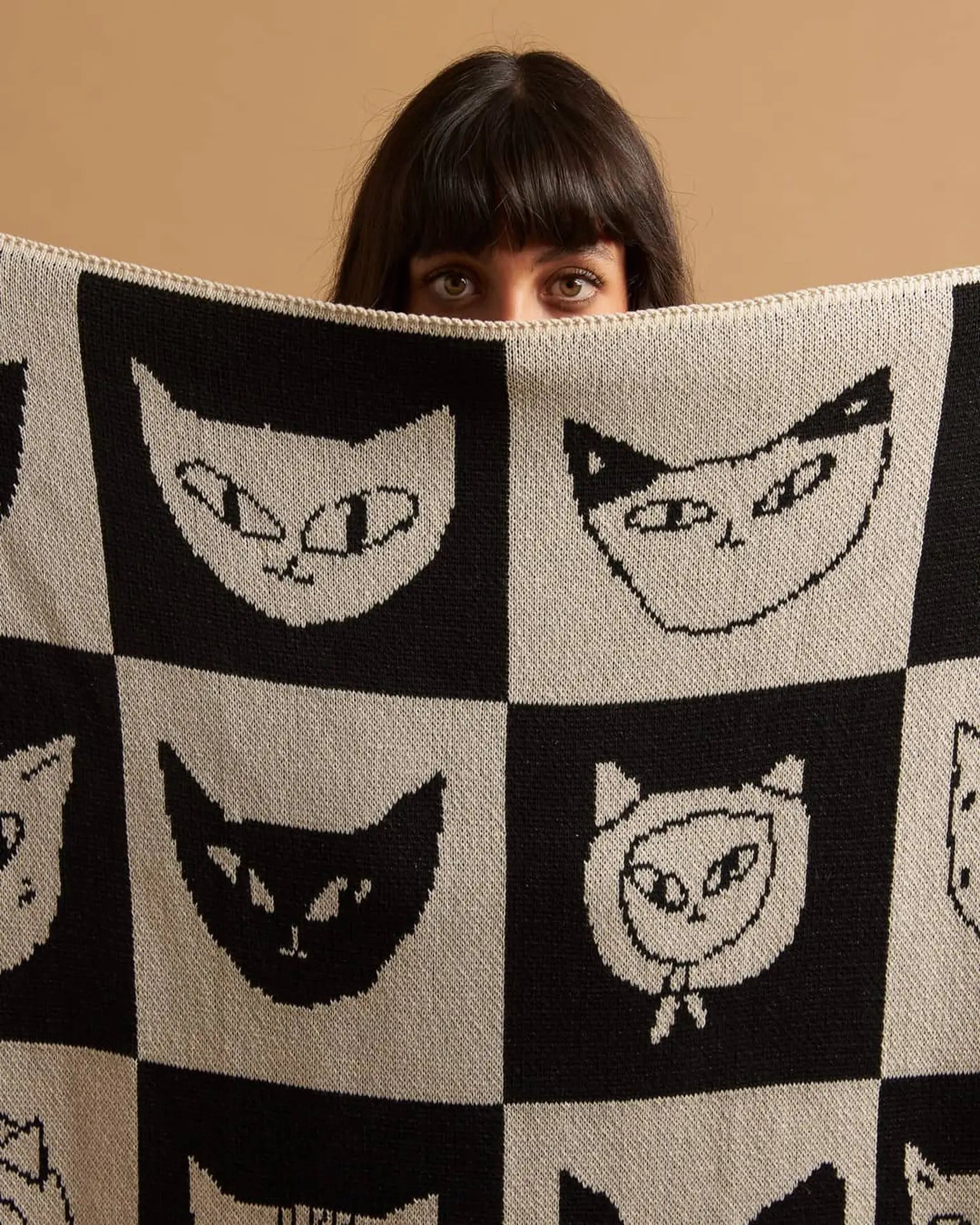 A person with dark hair and bangs peers over the Slowdown Studio Cat Bingo Mini Blanket, featuring a grid of stylized cat faces on a neutral background and crafted from a recycled cotton blend.