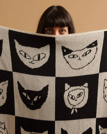 A person with dark hair and bangs peers over the Slowdown Studio Cat Bingo Mini Blanket, featuring a grid of stylized cat faces on a neutral background and crafted from a recycled cotton blend.