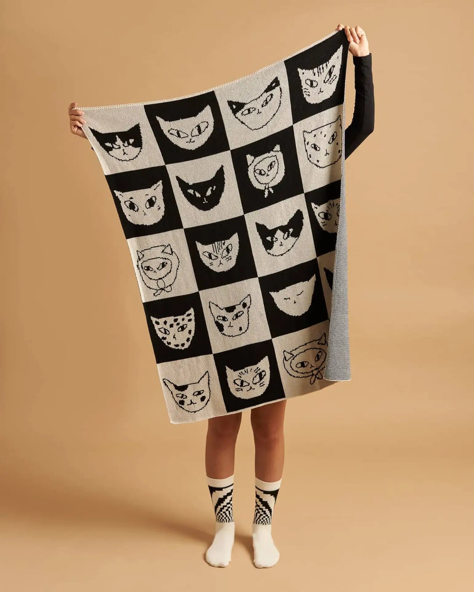 Someone holds the Slowdown Studio Cat Bingo Mini Blanket, which features a black-and-white checkerboard and cartoon cat faces. Made from recycled cotton blend, their socks also match the blanket’s cat design. Only legs and arms are shown.
