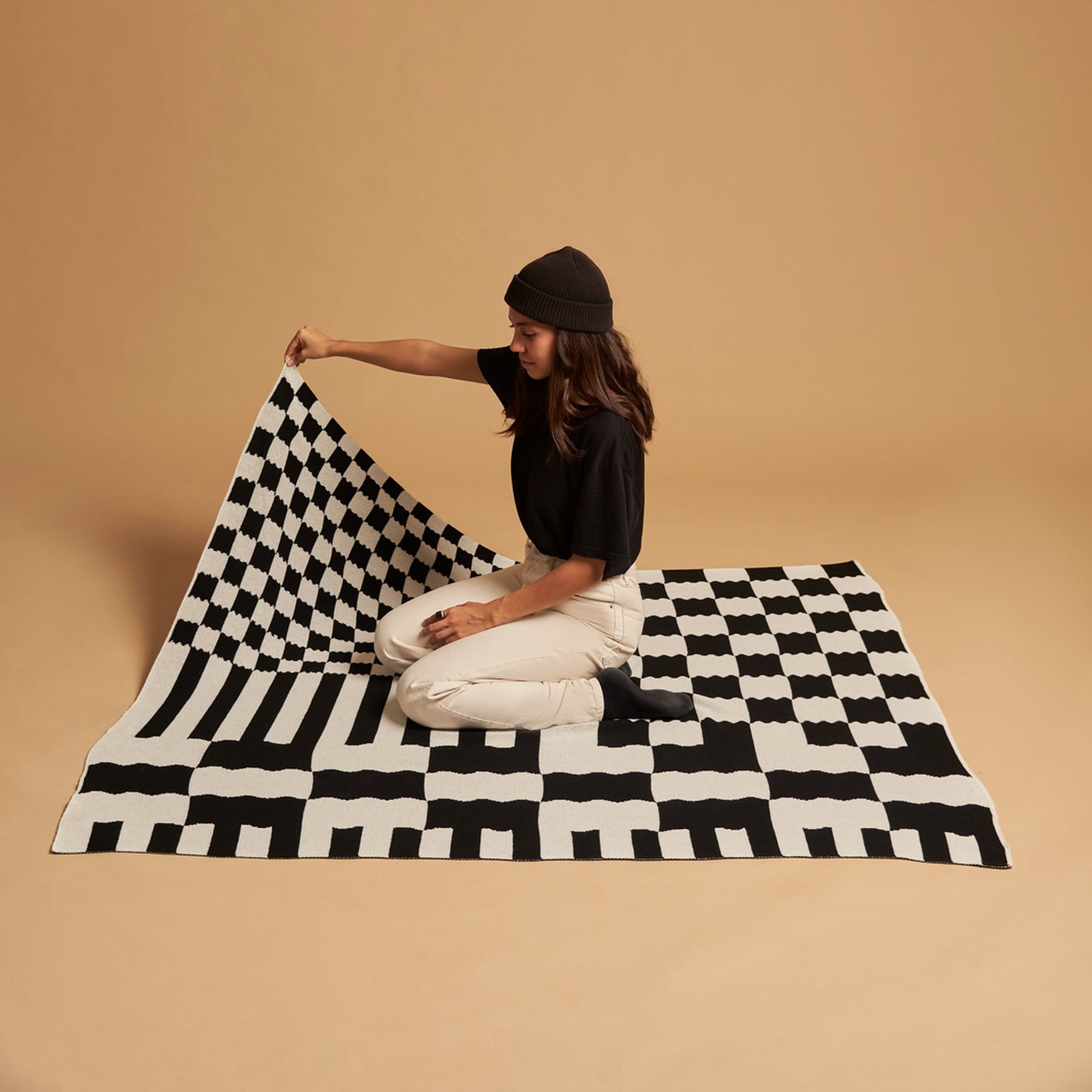 A person in a black beanie, black shirt, and light pants kneels on the Slowdown Studio Chilton Black/White Knit Blanket, lifting one corner, with a plain beige background behind them.