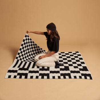 A person in a black beanie, black shirt, and light pants kneels on the Slowdown Studio Chilton Black/White Knit Blanket, lifting one corner, with a plain beige background behind them.