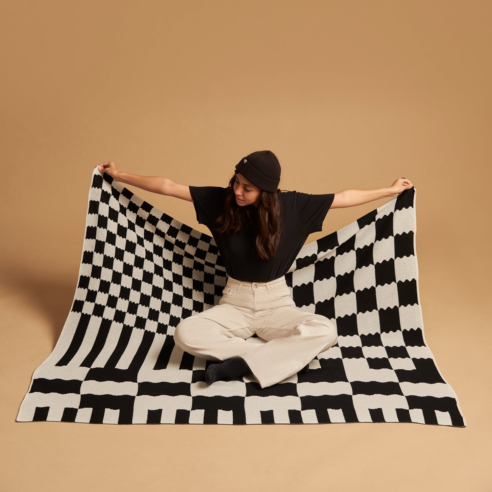 A person in a black beanie, black shirt, and light pants sits on the floor against a plain beige background, holding out the Slowdown Studio Chilton Black/White Knit Blanket by Slowdown Studio, featuring bold black and white checks.