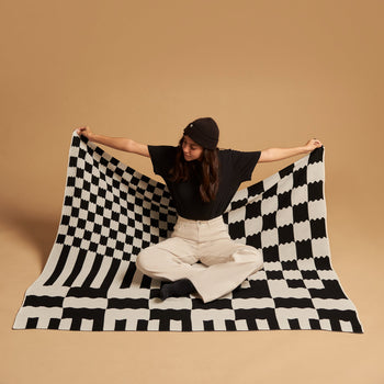 A person in a black beanie, black shirt, and light pants sits on the floor against a plain beige background, holding out the Slowdown Studio Chilton Black/White Knit Blanket by Slowdown Studio, featuring bold black and white checks.