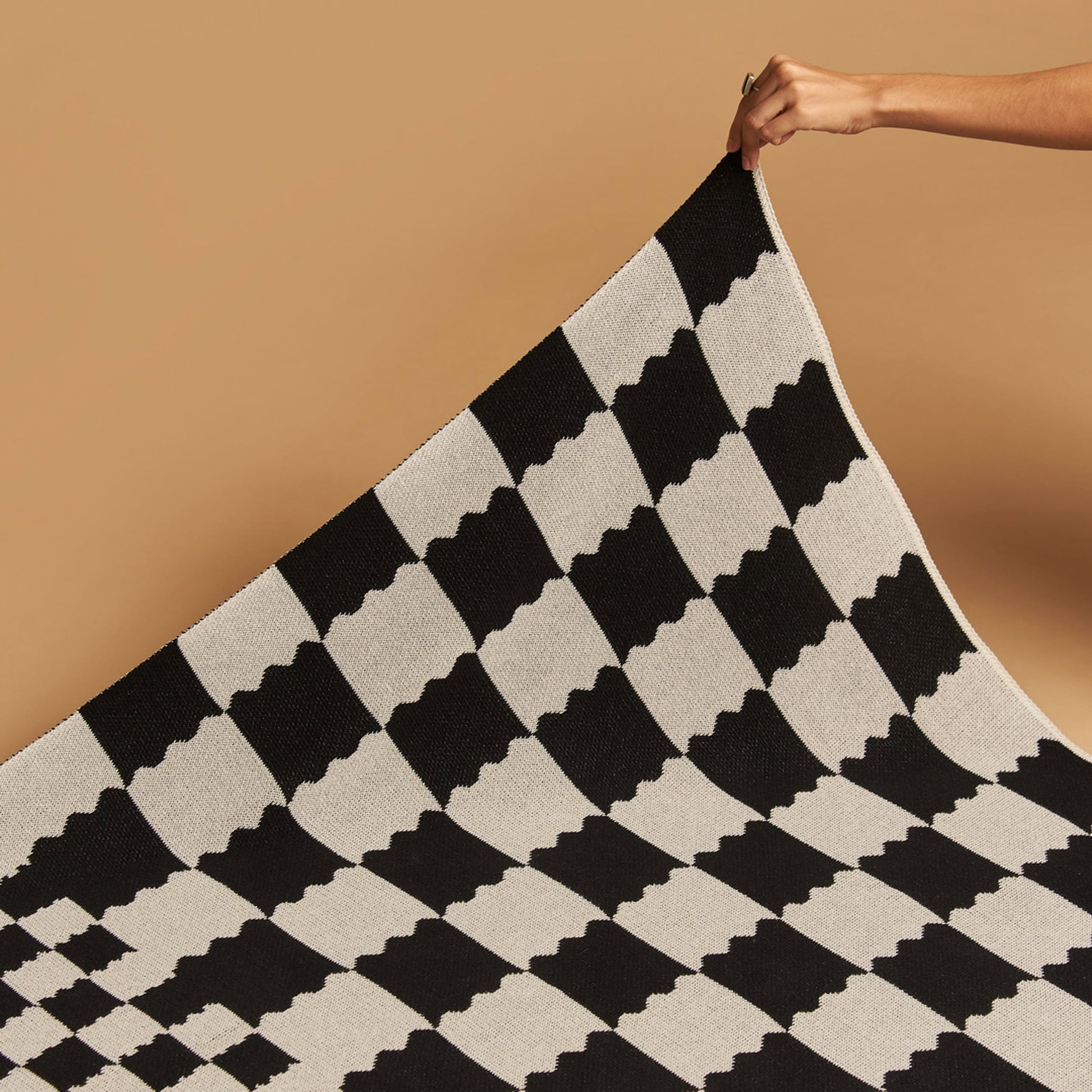 A hand lifts the corner of the Slowdown Studio Chilton Black/White Knit Blanket, made from recycled cotton with a black and white optical illusion checkered design, set against a plain beige background.