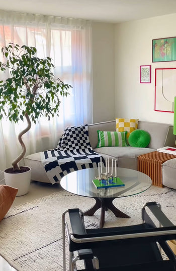A bright living room with a white sectional sofa, the Slowdown Studio Chilton Black/White Knit Blanket, colorful pillows and throws, a large potted tree, round glass coffee table, black chair, modern art on light walls, and sheer curtains.