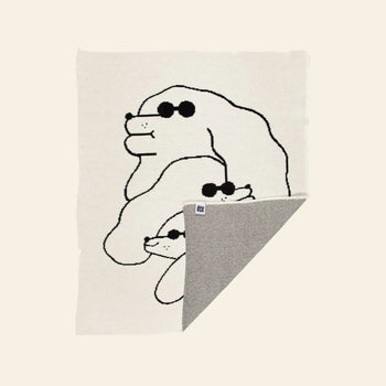 The Slowdown Studio Dawgs Mini Blanket is a cream-colored recycled cotton throw featuring a black line drawing of three dogs in sunglasses, stacked by size, with a folded corner showing the contrasting gray underside.