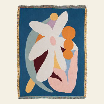 The Slowdown Studio Delaney Throw by Slowdown Studio is an eco-friendly woven blanket made from recycled cotton, featuring multicolored abstract shapes, a large white flower, orange circles, pastel accents, and vibrant fringed edges on deep blue.