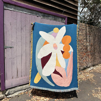 The Slowdown Studio Delaney Throw, made from recycled cotton with an abstract floral design, hangs outdoors before a weathered purple wooden door and brick wall, dry leaves scattered on the ground.