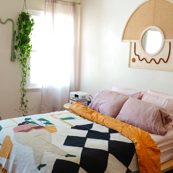 A cozy bedroom with soft pink pillows, the Slowdown Studio Dresden Throw by Slowdown Studio—a colorful geometric quilt crafted from recycled cotton—plus a mustard yellow blanket, hanging plant, sheer curtains, and a rattan wall mirror above the bed.