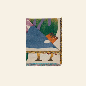 The Slowdown Studio Dumont Throw features colorful abstract patterns in blue, green, pink, orange, and yellow with a multicolored fringe. Made from recycled cotton, it’s shown folded and stacked on a light background.