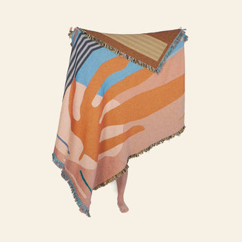 A person displays the Slowdown Studio Hazlewood Throw, a large, eco-friendly blanket with orange, blue, pink, brown, and beige abstract designs; only their feet peek out beneath this sustainable Slowdown Studio statement piece.