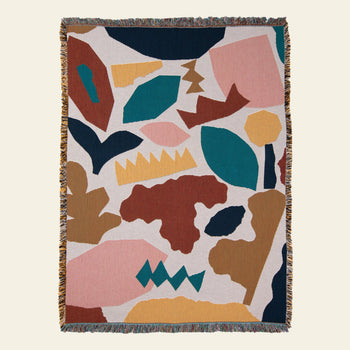 The Slowdown Studio Maple Throw is a fringed woven blanket made from recycled cotton, featuring an abstract pattern in teal, mustard yellow, rust brown, pink, and dark blue on a light background.