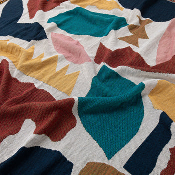 The Slowdown Studio Maple Throw by Slowdown Studio features a colorful abstract pattern in teal, mustard yellow, burgundy, pink, and navy on white. Made from recycled cotton, its textured surface and soft wrinkles add unique character.
