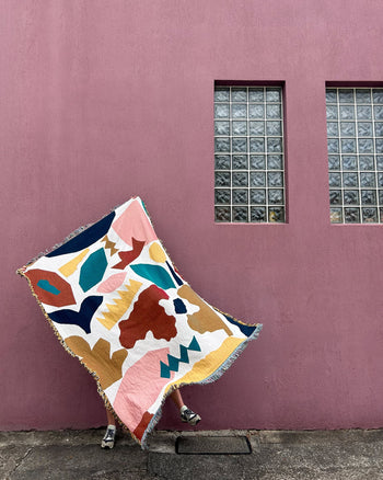 A person displays the Slowdown Studio Maple Throw, a colorful abstract-patterned blanket crafted from recycled cotton by Slowdown Studio, in front of a mauve wall. Only their feet in sandals peek out below this eco-friendly throw.