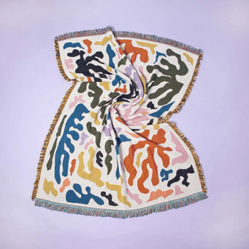 The Slowdown Studio Mercer Throw is a multicolored woven blanket made from recycled cotton, featuring abstract wavy patterns and fringed edges, twisted in the center and laid flat on a pale lavender surface.