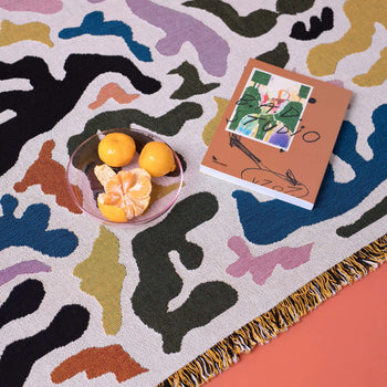 The Slowdown Studio Mercer Throw, featuring a vibrant abstract pattern and made from recycled cotton, serves as a backdrop for a glass bowl of tangerines and an illustrated book resting on the fringed rug.