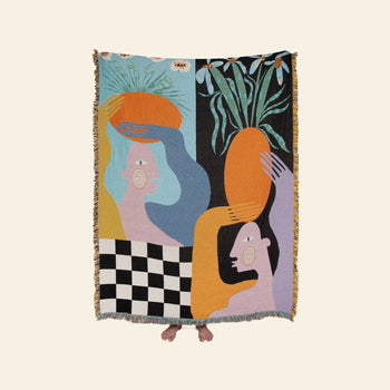 A person holds the Slowdown Studio Moretti Throw by Slowdown Studio, an eco-friendly tapestry made from recycled cotton with abstract colorful figures, large orange shapes, a checkerboard base, and leafy plants. Only their toes peek out.