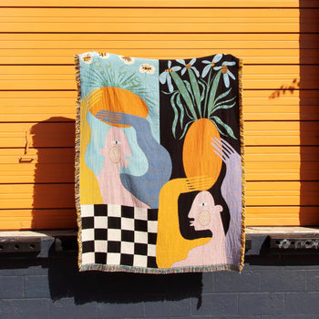 The Slowdown Studio Moretti Throw by Slowdown Studio, made from recycled cotton and featuring abstract human figures, flowers, and a black-and-white checkerboard design, hangs against a bright orange wall.