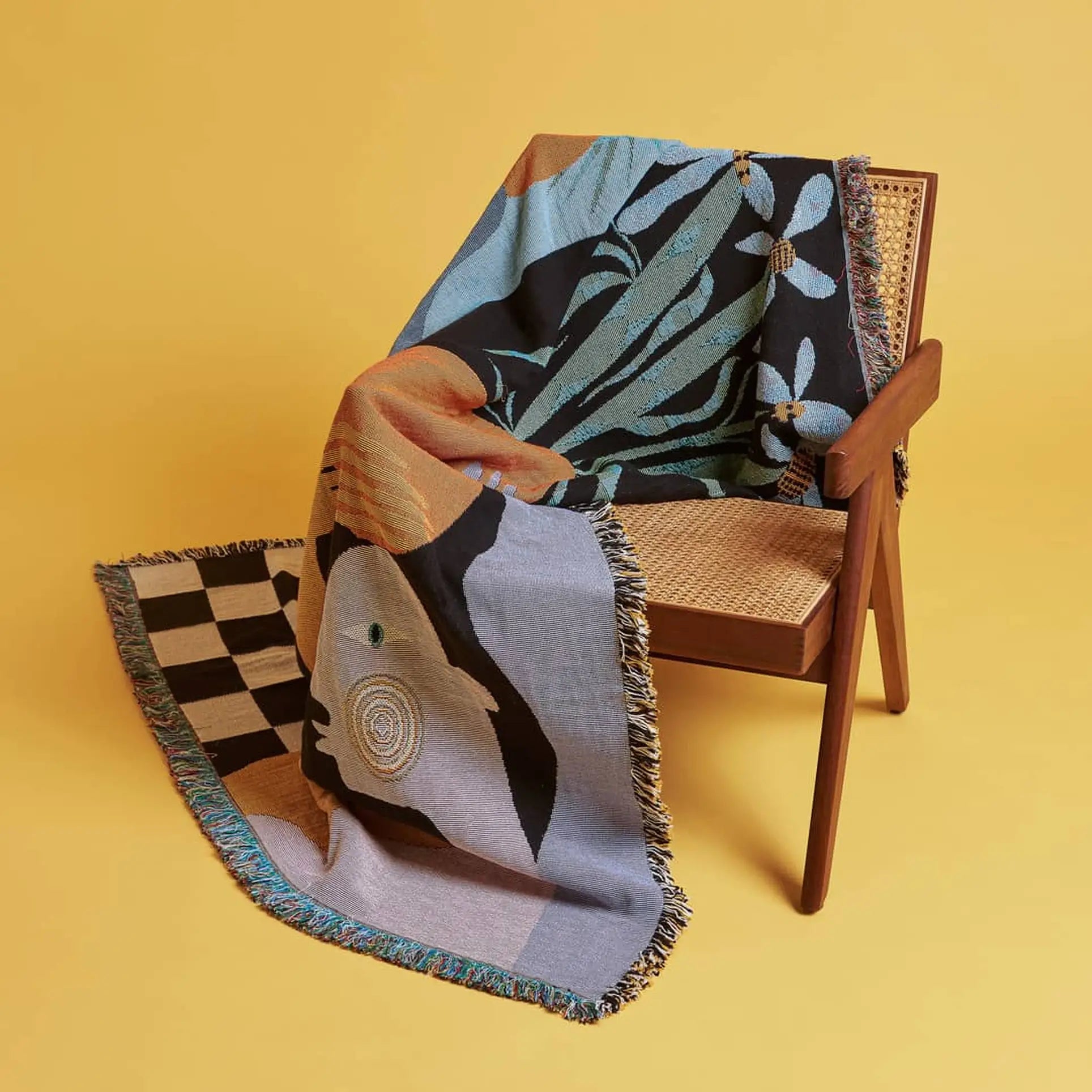 The Slowdown Studio Moretti Throw by Slowdown Studio, featuring abstract patterns and fringe and made from recycled cotton, is draped over a wooden cane-backed chair set against a plain yellow background.