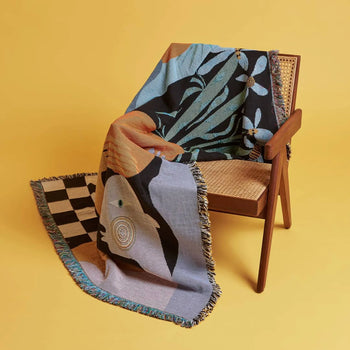 The Slowdown Studio Moretti Throw by Slowdown Studio, featuring abstract patterns and fringe and made from recycled cotton, is draped over a wooden cane-backed chair set against a plain yellow background.