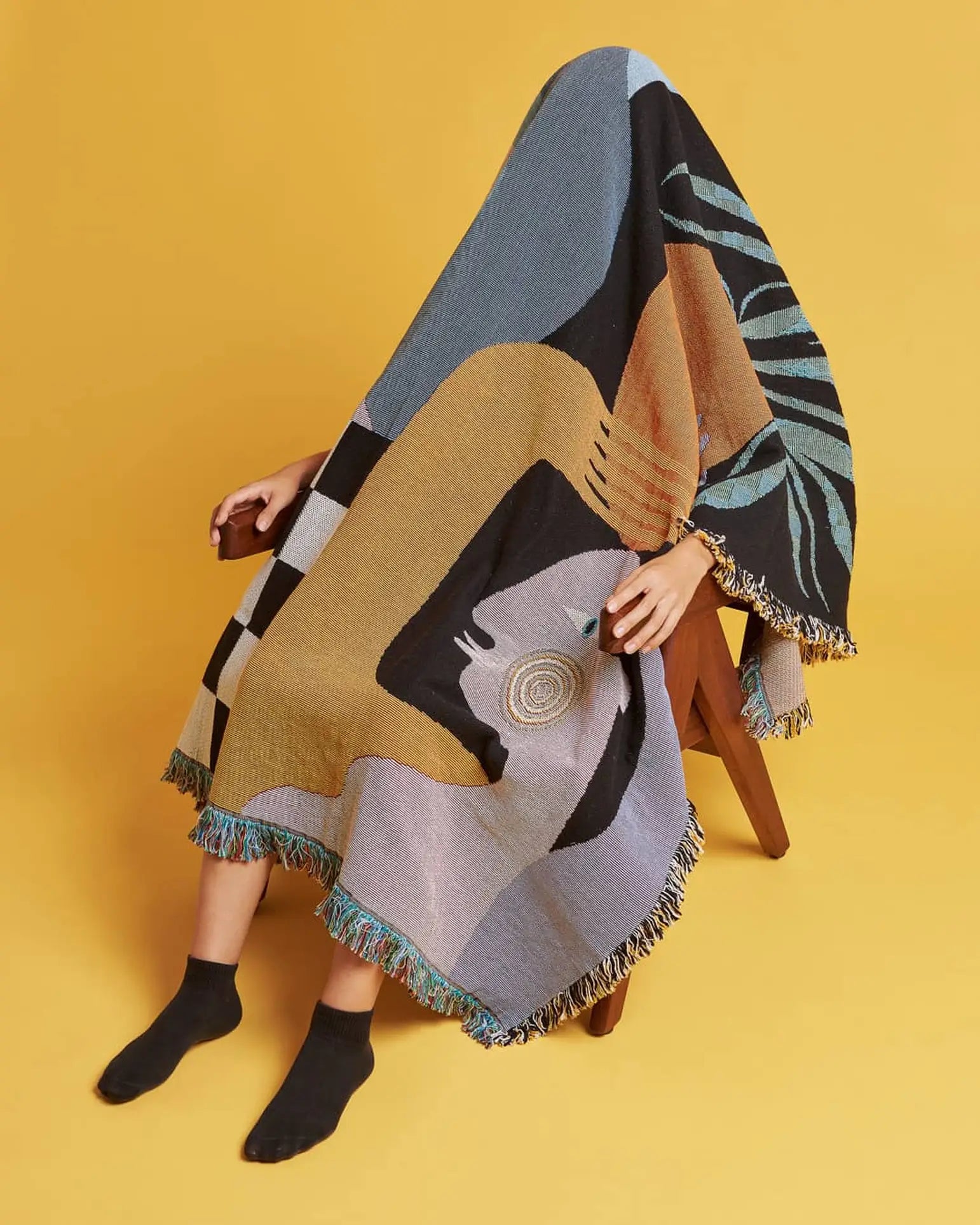 A person sits on a wooden chair wrapped in the colorful, abstract-patterned Slowdown Studio Moretti Throw by Slowdown Studio, made from recycled cotton. Only their hands and lower legs in black socks show against a solid yellow background.