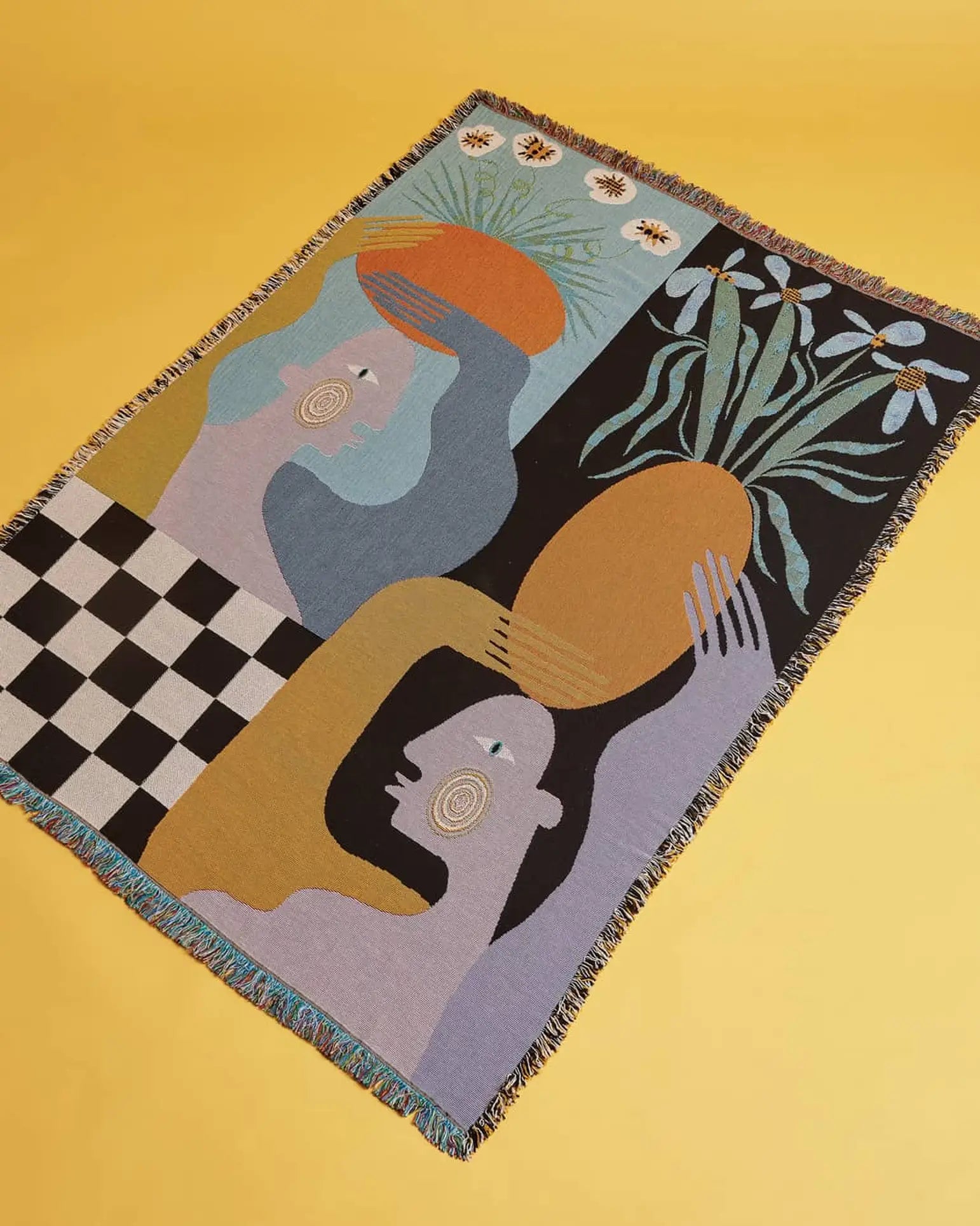 The Slowdown Studio Moretti Throw by Slowdown Studio is a woven tapestry made from eco-friendly recycled cotton, showcasing abstract human faces, floral motifs, checkerboard patterns, and bold muted shapes on a yellow background.