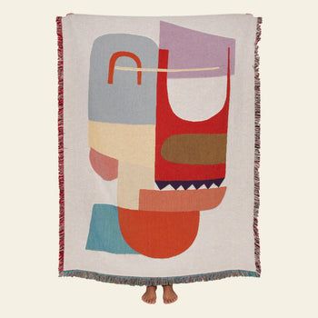 A person holds up the Slowdown Studio Oroza Throw, a large, colorful woven blanket by Slowdown Studio made from recycled cotton, showing its abstract geometric design with their feet visible below the edge.