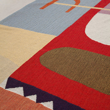 A close-up of the Slowdown Studio Oroza Throw, a colorful woven textile by Slowdown Studio made from recycled cotton, featuring geometric shapes in red, blue, beige, and olive green with zigzag and curved patterns.