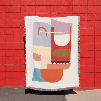 The Slowdown Studio Oroza Throw by Slowdown Studio, made from recycled cotton and featuring a colorful geometric pattern, hangs against a vivid red brick wall, with part of it casting a shadow on the sidewalk.