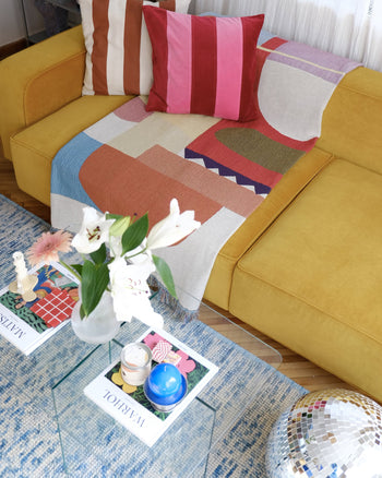 A yellow sofa features colorful, geometric pillows and the eco-friendly Slowdown Studio Oroza Throw made from recycled cotton. In front, a glass coffee table displays magazines, candles, a vase with white lilies, and a disco ball sits nearby.