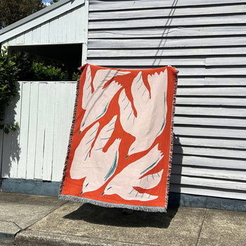 The Slowdown Studio Passaro Throw by Slowdown Studio is made from recycled cotton and features abstract red, white, and teal shapes. It hangs on a light gray wooden wall outside, casting a shadow on the sidewalk in bright sunlight.