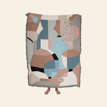 A person stands holding up the Slowdown Studio Pollard Throw by Slowdown Studio, an eco-friendly blanket crafted from recycled cotton, featuring an abstract geometric design in muted blue, beige, pink, black, and gray. Only their bare feet show below.