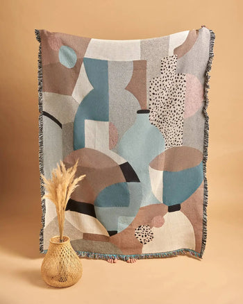 A person holds up the Slowdown Studio Pollard Throw, a large recycled cotton blanket in muted blue, brown, and beige hues. In front, an eco-friendly woven vase with pampas grass sits against a beige background. Only toes peek beneath the throw.