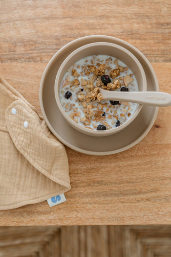 A beige bowl of granola with milk and dried berries sits on a matching plate, paired with a Soft Spot Soft Baby Spoon for easy feeding. Next to it is a beige cloth bib on a wooden surface, perfect for little ones learning to eat.